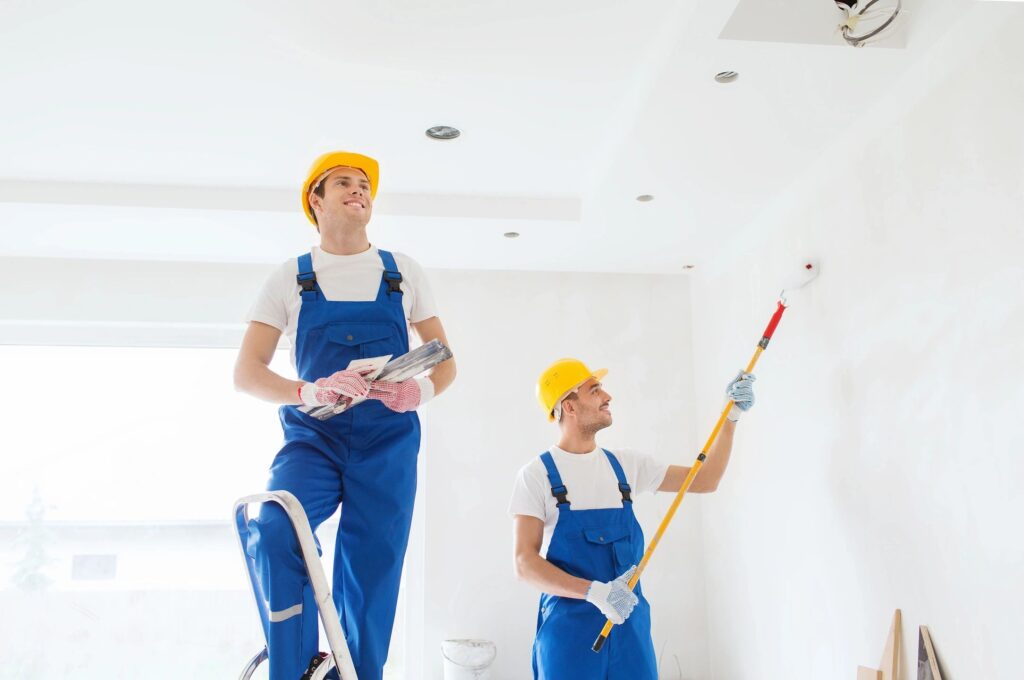 Two people in blue overalls and hard hats are painting a room.